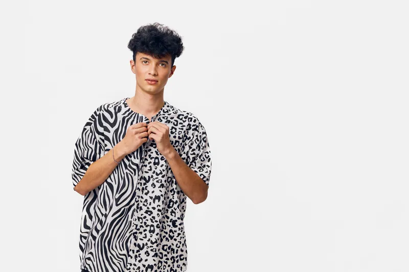 young man wearing black and white animal print shirt stands against plain white background, casual style with curly hair and relaxed expression, contemporary fashion portrait