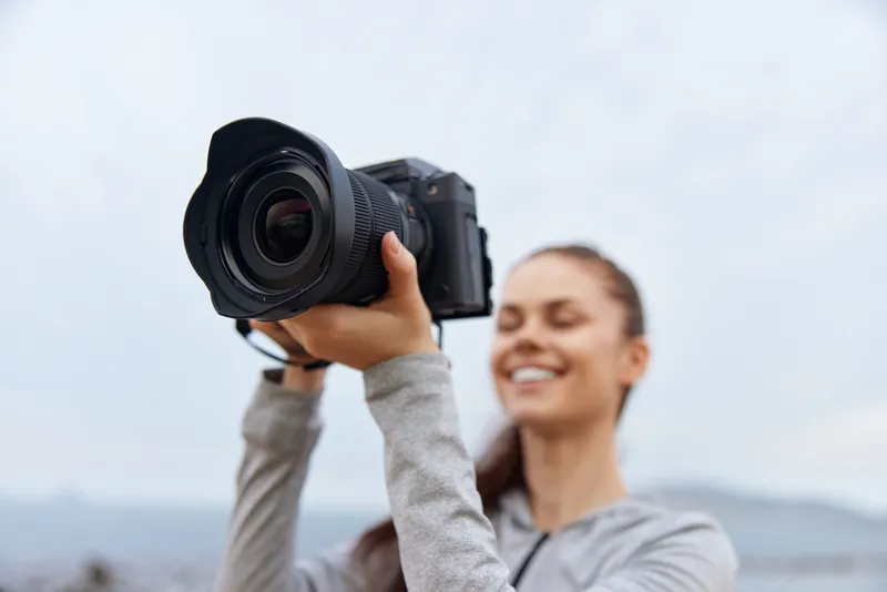 Happy young woman holding a camera outdoors, showcasing a passion for photography with a smile against a cloudy background, perfect for lifestyle and travel concepts
