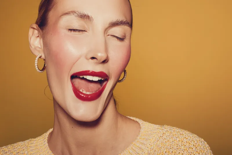 Close-up of a smiling woman with red lipstick and hoop earrings winking with eyes closed. Warm yellow background and soft lighting create a joyful and playful mood in the portrait.