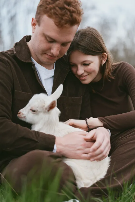 A couple sits outdoors, cradling a white goat kid in their arms, a tender, candid moment of connection. A lifestyle image highlighting companionship, warmth, and gentle care between humans and
