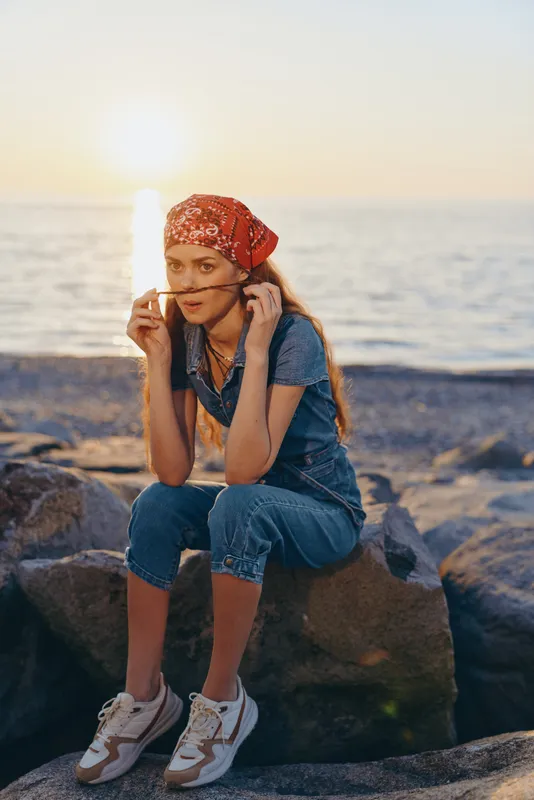lifestyle woman wearing modern boho-western denim street style outfit with red bandana, seated on rocks by the ocean during golden hour in warm film color tones