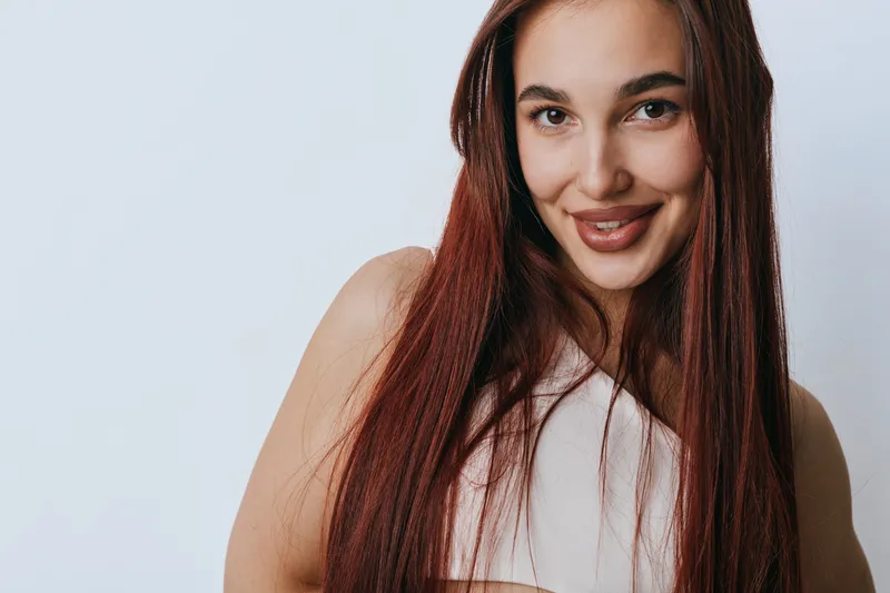 Young woman with long red hair smiling and looking at camera in studio. Portrait of cheerful female with natural makeup and casual outfit on light background.