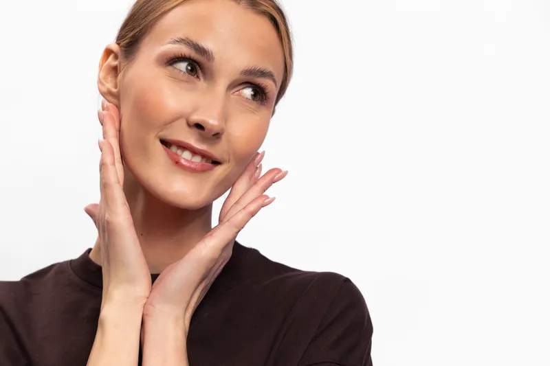 Smiling woman with clear skin touches her face gently, looking away on white background. Concept of skincare, beauty, self-care, and facial wellness with natural expression.