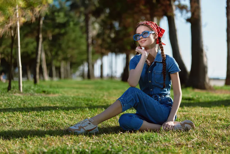 lifestyle woman in modern boho-western denim street style sitting on grass with sunglasses and red bandana, natural light, film color aesthetics
