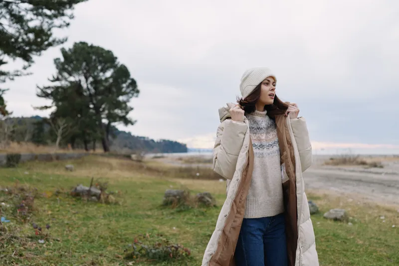 Woman in a long beige winter coat knit sweater and beanie standing in an open field by a lake under a pale sky enjoying chilly weather and calm landscape