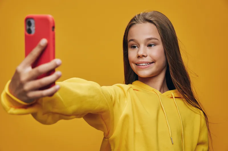 Teenage girl in yellow hoodie smiling and taking selfie with red smartphone on yellow background. Long hair, casual style and cheerful expression for social media post or communication.