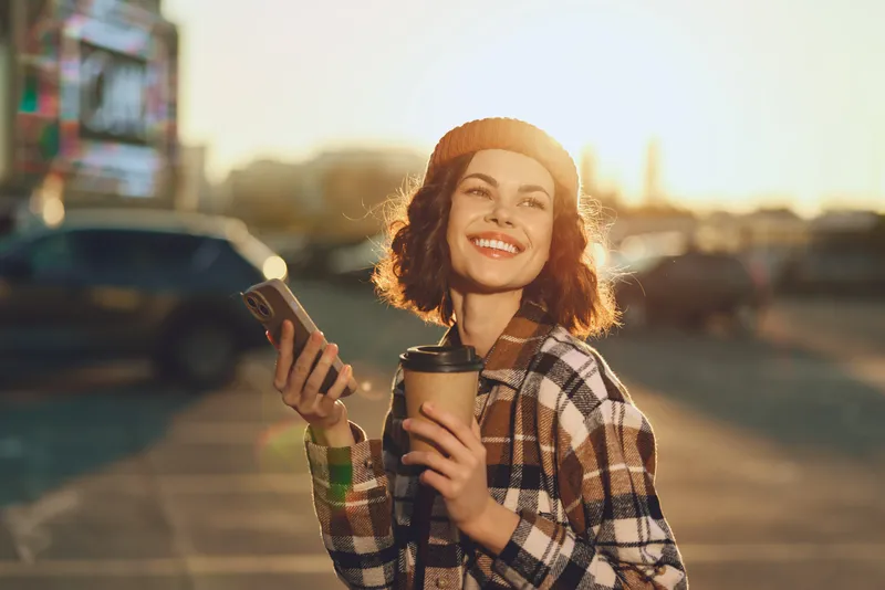 Woman holding coffee and smartphone, smiling in golden hour urban street, candid beanie portrait conveying authenticity, mindful living and emotional storytelling with warm glow.