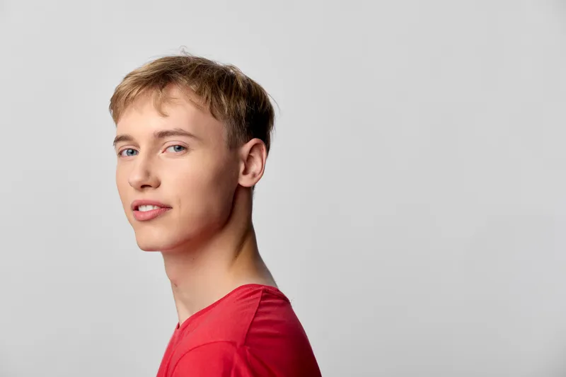 Young man in a red shirt stands in a studio with a calm expression, posing against a neutral background. Clean lighting highlights facial features and style, conveying casual confidence and modern