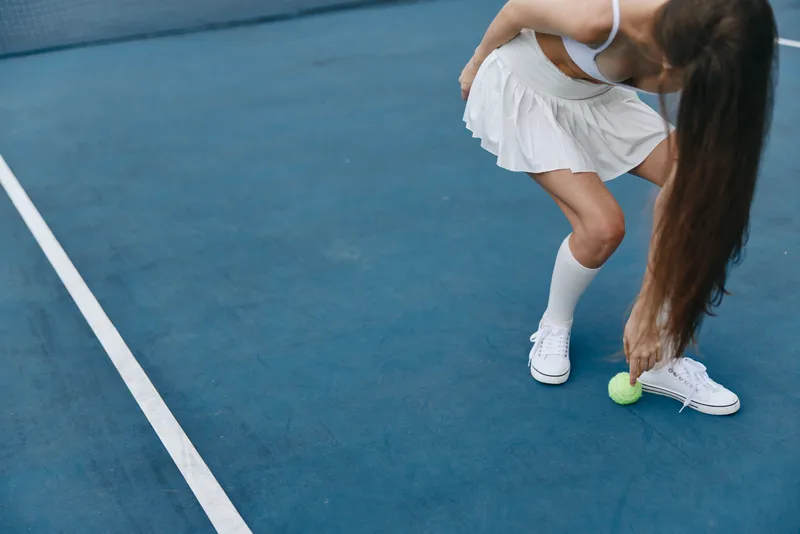 Young female tennis player in a sporty outfit bends to pick up a tennis ball on a blue court, showcasing determination and athleticism during a sunny day