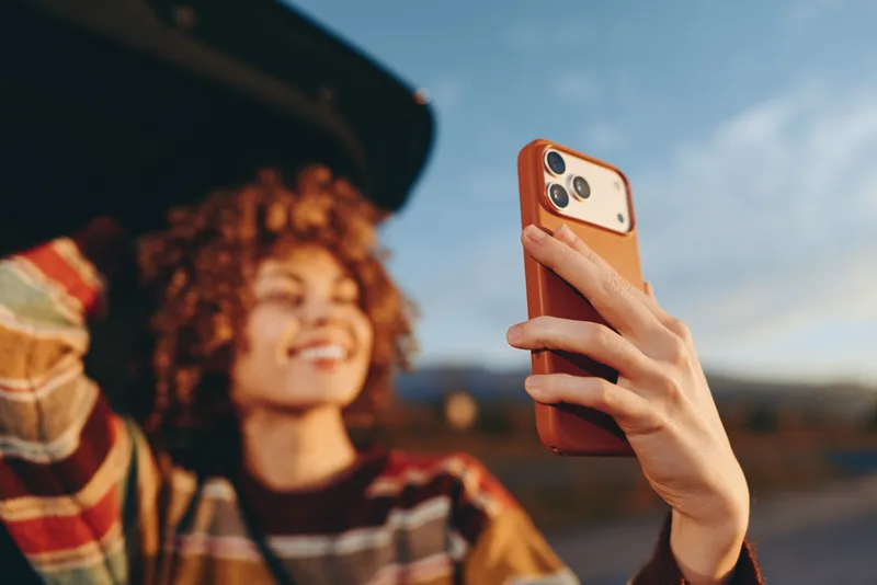 Woman with curly hair takes a selfie outdoors, holding a smartphone in her hand against a scenic sky. Lifestyle moment of everyday technology, warm daylight, travel mood.