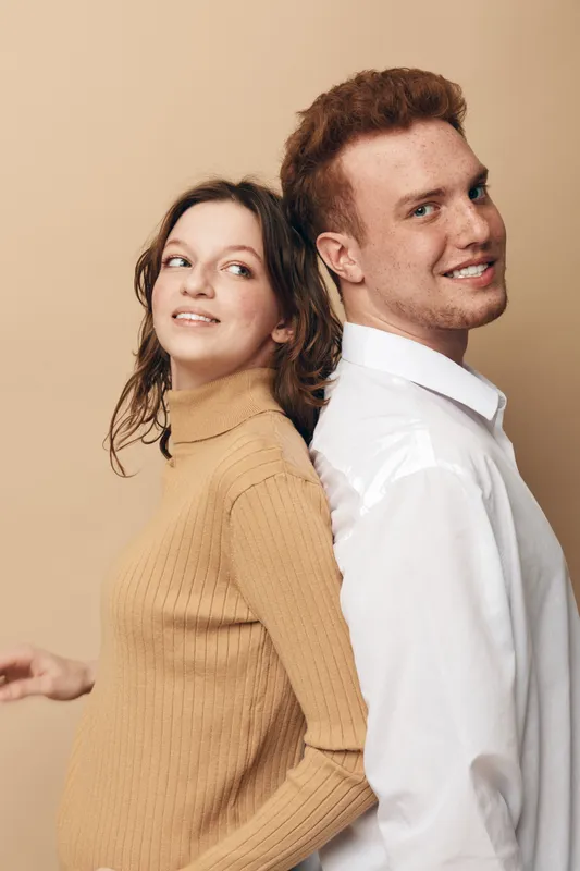 Young man and woman stand back to back smiling, wearing casual clothes on beige background. Concept of friendship, relationship, youth, and positive emotions in studio portrait.