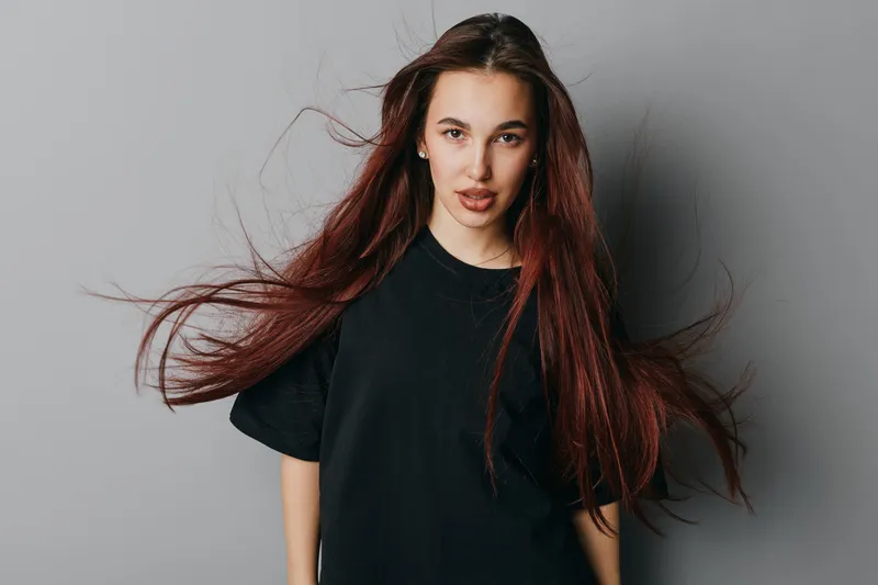 Young woman with long red hair wearing black t shirt standing against plain gray background looking at camera with confident expression and natural makeup indoors.