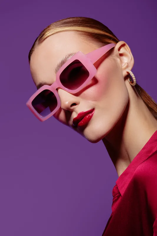 Stylish woman with pink sunglasses and red lipstick poses against purple background. Fashion portrait with bold makeup and modern accessories. Concept of glamour and confident beauty.