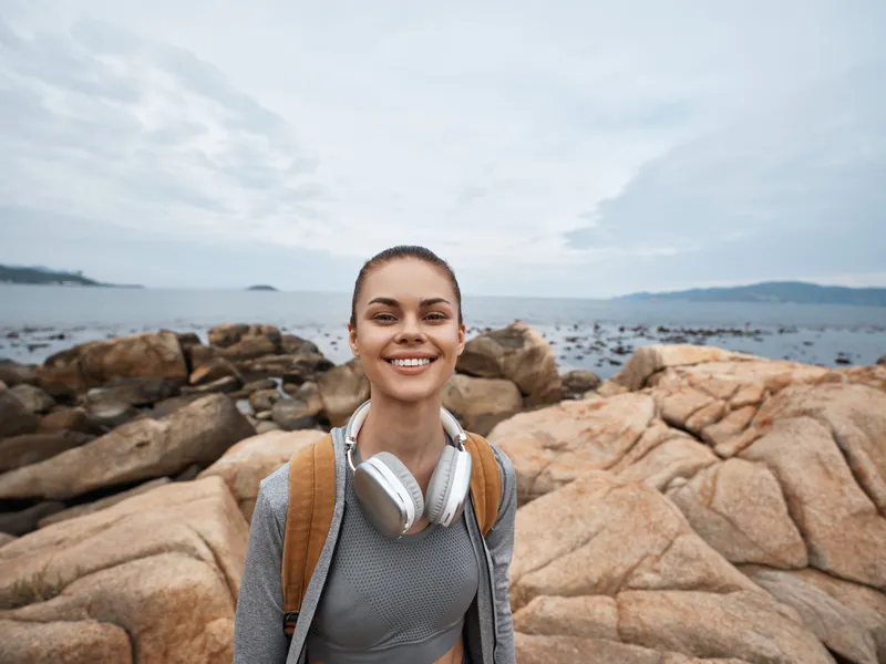 Young woman smiling by the sea, wearing headphones and sportswear, showcasing energy and joy in a coastal outdoor setting