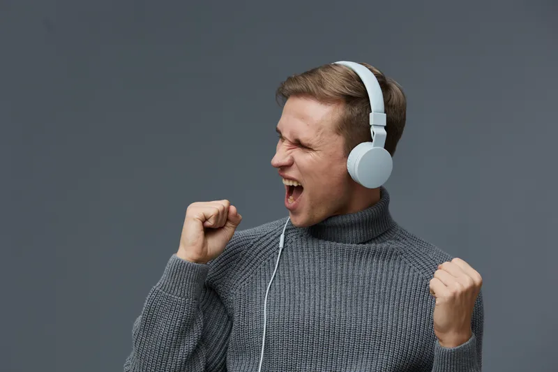 Young man with short brown hair wearing gray sweater and white headphones expressing excitement and joy, clenched fists and open mouth, studio portrait on solid gray background, lifestyle celebration 