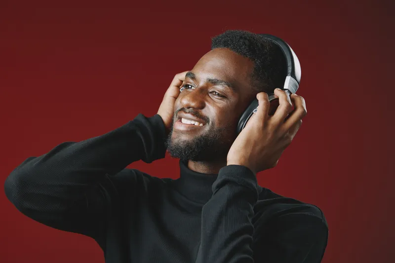 Joyful young man enjoying music with headphones against a red background, wearing a black turtleneck, expressing happiness and relaxation