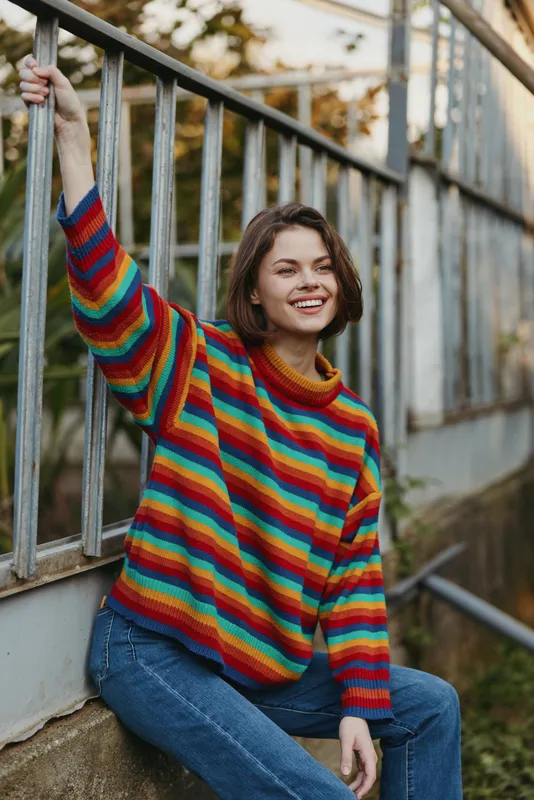 Young woman in her 20s, striped sweater, smiling and leaning on metal railing while sitting on urban stairs, casual outfit, short brown hair, relaxed autumn lifestyle portrait.