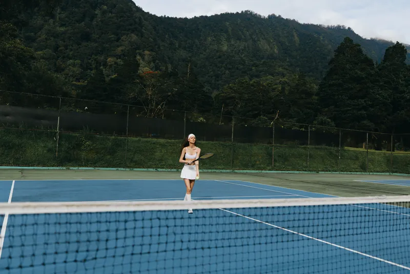 Young female tennis player in white sportswear smiling and playing on a blue court surrounded by green mountains, creating an energetic and vibrant atmosphere