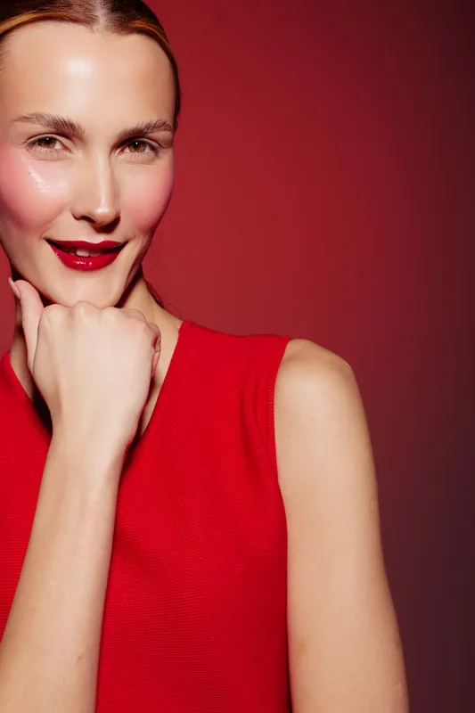 Confident woman in red dress poses with hand near chin against red background. Studio portrait with bright makeup and smooth skin. Concept of beauty, fashion, and elegance.