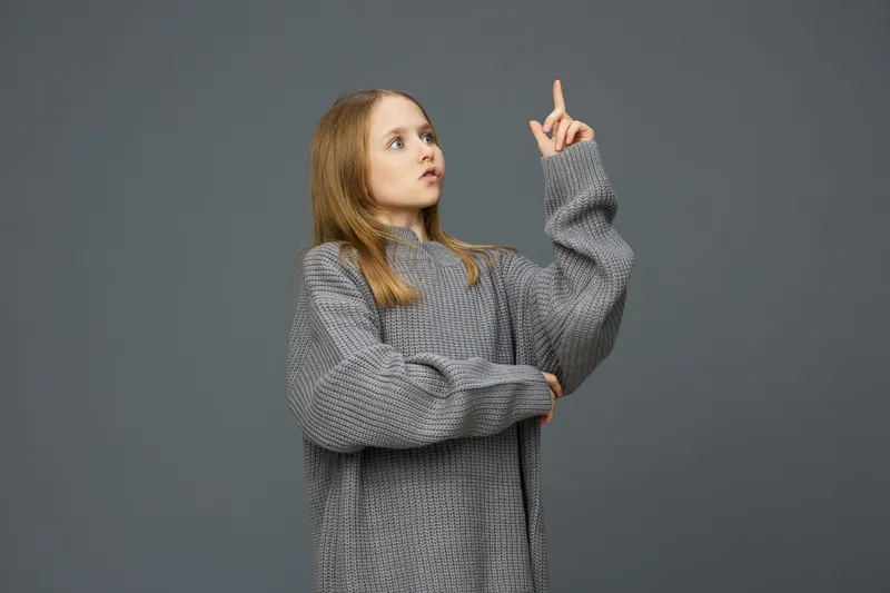 Young girl in an oversized gray sweater pointing upward, displaying curiosity and engagement against a neutral gray background Perfect for concepts of inspiration and creativity