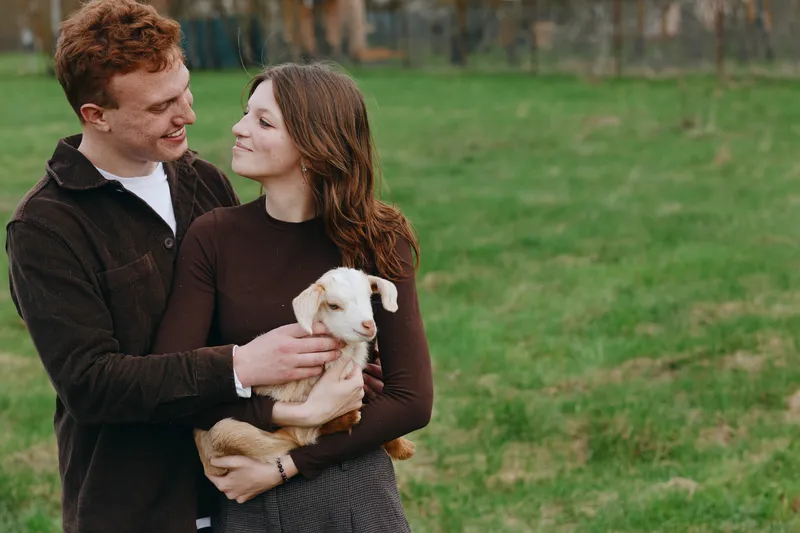 A young couple embraces outdoors as the woman holds a small goat in her arms, sharing a tender moment in a green open field during a casual lifestyle shoot.