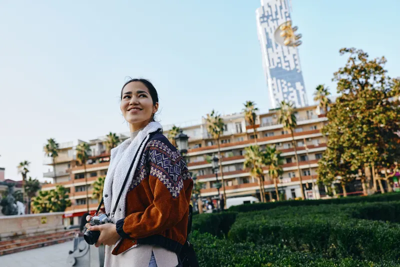 Woman with camera outdoor urban setting, smiling beside architecture and palm trees, capturing travel lifestyle and candid moments in daylight, street scene with greenery and modern buildings,