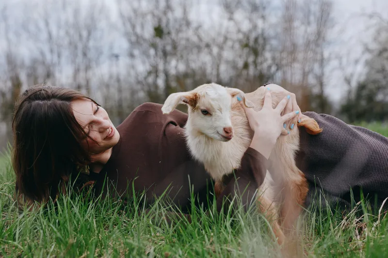 Cheerful woman outdoors lying in grass, gentle interaction with a curious goat kid, warm light, relaxed mood, candid countryside lifestyle, soft breeze, natural colors, tranquil scene