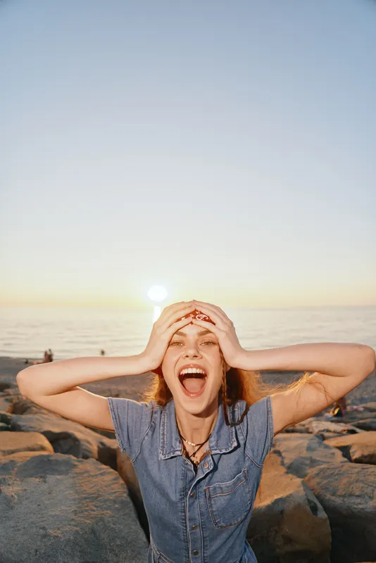joyful woman wearing modern boho-western denim street style laughing outdoors at sunset on rocky beach with warm film color tone and carefree vibe