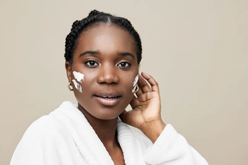 Young woman with skincare cream on her face, wearing a bathrobe, expressing joy, against a neutral background, promoting healthy skin and beauty care
