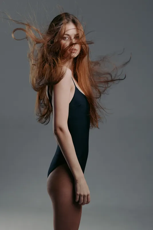 Woman with long hair in black swimsuit posing against gray background, movement, elegance, beauty