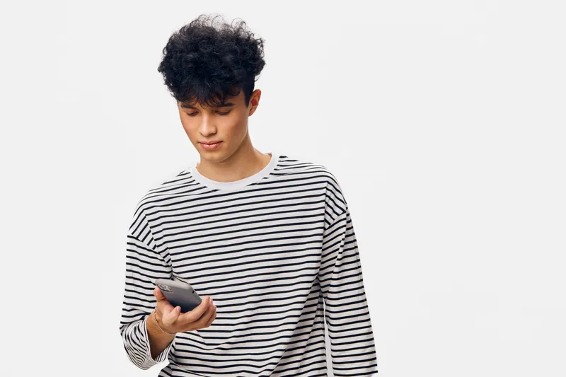 young man in striped shirt looking at smartphone with casual and thoughtful expression on white background, modern lifestyle technology concept