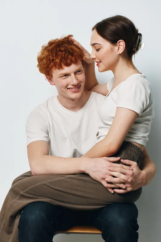 Couple, young man and woman in casual clothes sitting together, smiling and showing affection, happiness and connection in studio with white background and cozy atmosphere.