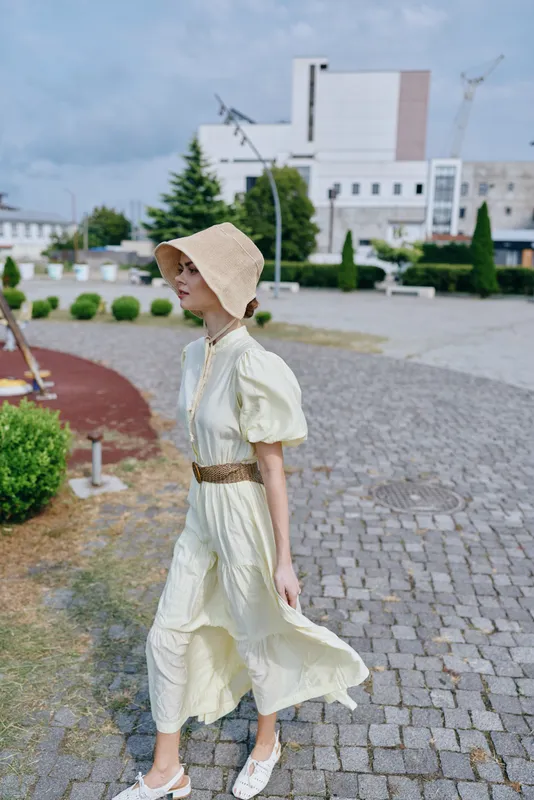 woman in light yellow dress and wide brim hat walking on a cobblestone path outdoors with modern buildings in the background, a sunny summery fashion scene with graceful movement