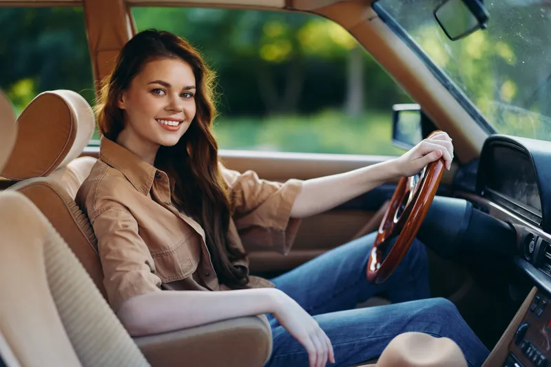 Young woman smiling joyfully in a vintage car, wearing casual attire, creating an inviting atmosphere with natural background and warm colors.