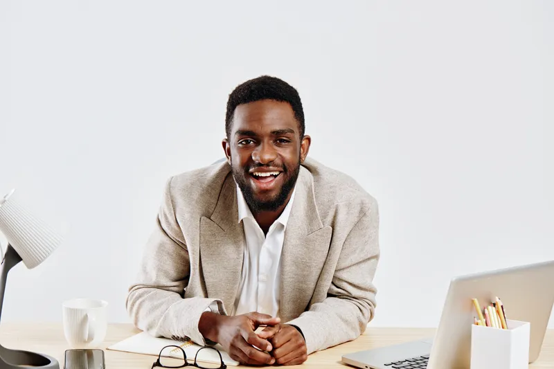 Smiling young African American man in beige blazer and white shirt sitting at desk with laptop in modern office environment, joyful expression, professional mood, workspace setup, casual business styl