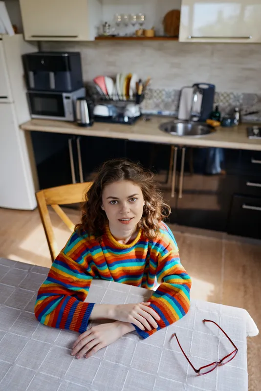 A woman with a colorful striped sweater sits at a table in a bright kitchen, looking at the camera. A cozy home lifestyle portrait conveying warmth, casual mood, and comfortable everyday life.