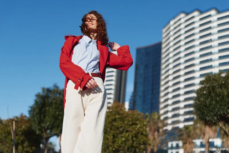 Fashionable woman with curly hair wearing a red jacket and white trousers stands outdoors on a city street, smiling under bright daylight, showcasing confidence and urban style and bold urban energy.