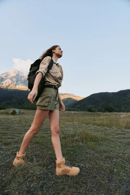 A young woman with a backpack hikes through a grassy mountain landscape wearing shorts and boots. The clear sky and distant peaks create a natural outdoor adventure scene.