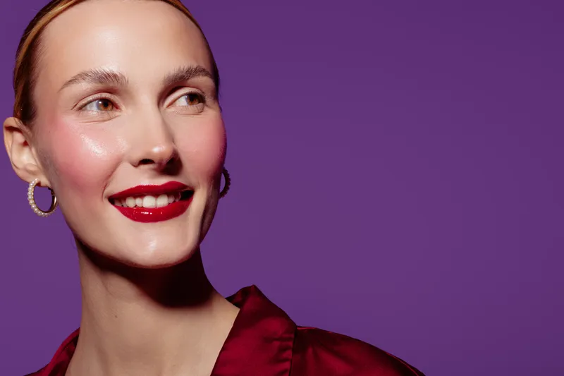 Smiling woman with red lipstick and hoop earrings looks away on purple background. Close-up portrait with natural skin glow and shiny makeup. Concept of beauty, fashion, and confidence.