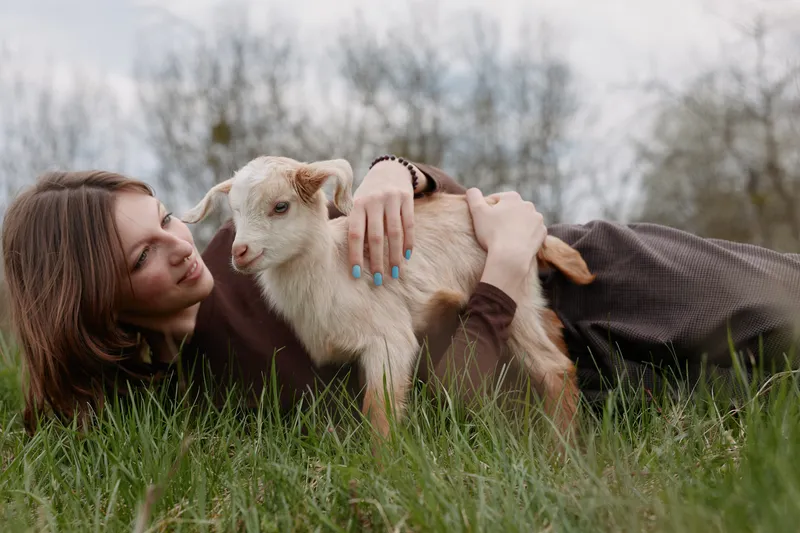 Young woman lying in grass outdoors, gently embracing a playful goat kid, capturing a warm moment of companionship, nature connection, soft light, relaxed mood, casual lifestyle.