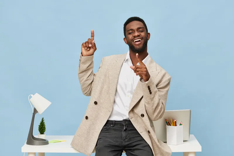 Cheerful happy young man with dark skin, stylish beige blazer and white shirt, showing thumbs up and smiling broadly, standing at modern desk with lamp, plant, and stationery, cheerful office vibe.