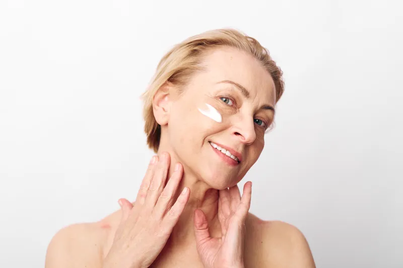 Smiling mature woman applies white face cream on cheek with hands near neck on white background. Concept of skincare, aging beauty, self-care, wellness and natural glow.