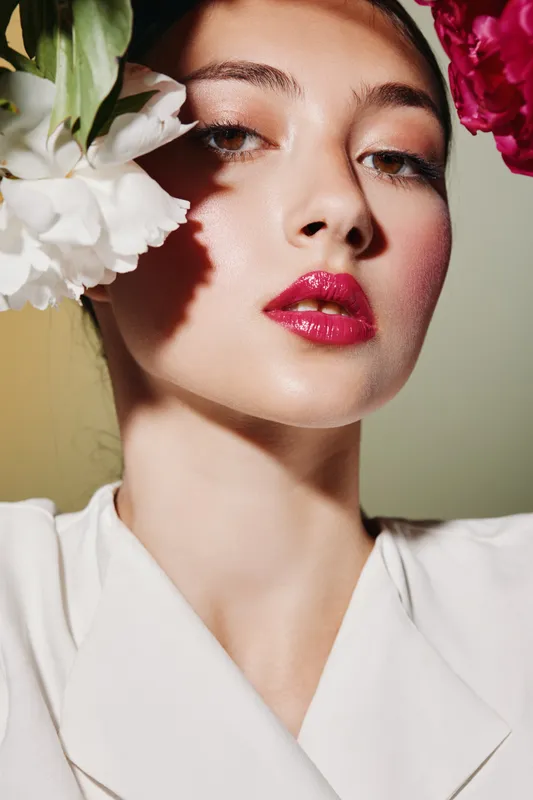 Close-up of a young woman with glossy pink lips and dewy complexion wearing a white top with vibrant floral elements around her face. Beauty concept for fashion and skincare.