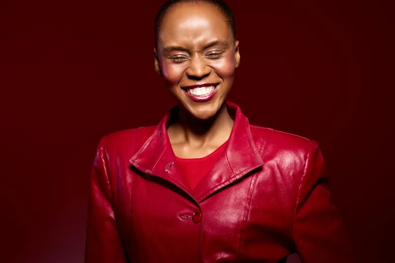 Happy African woman with dark skin wearing a red leather jacket smiles with closed eyes on a red background. Concept of joy, fashion, confidence, and vibrant style.