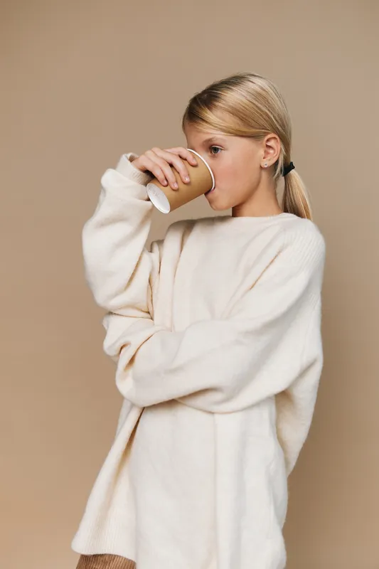 Young woman drinks from a paper cup in a cozy white sweater, posed against a soft beige background. Subtle lighting enhances texture, creating a calm casual lifestyle portrait.