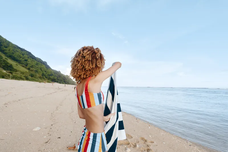 Woman on beach by sea coastline, summer travel mood, looking toward horizon, curly hair and colorful dress, carrying tote bag, warm sunlight, calm waves, natural colors