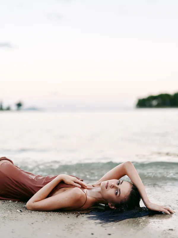 A serene beach scene features a relaxed woman resting on the sand by gentle waves, modeling casual eveningwear, soft light, tranquil mood, subtle elegance, coastal calm.