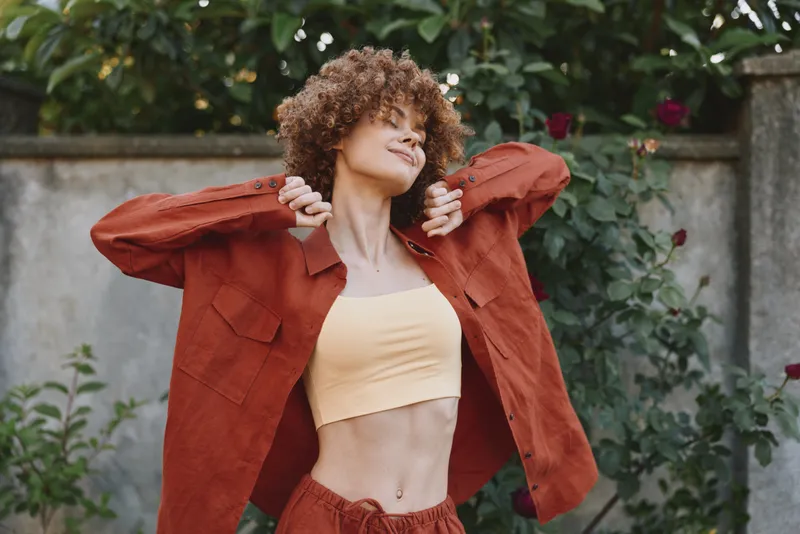 A woman wearing a rust jacket over a pale yellow crop top poses outdoors, arms lifted, smiling with ease. A street lifestyle concept emphasizing casual fashion, confidence, and daily self expression