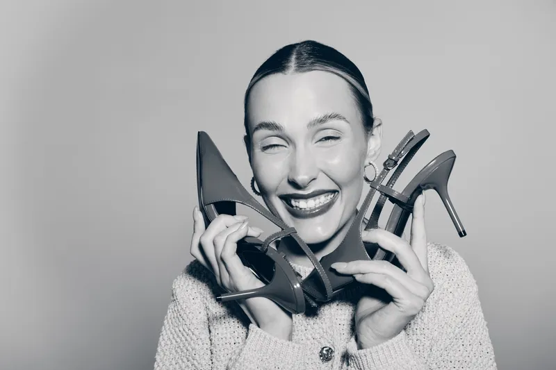 Happy caucasian woman with fair skin and slicked hair smiles broadly holding two high heel shoes near her face on plain background. Concept of fashion, style, joy, and footwear.