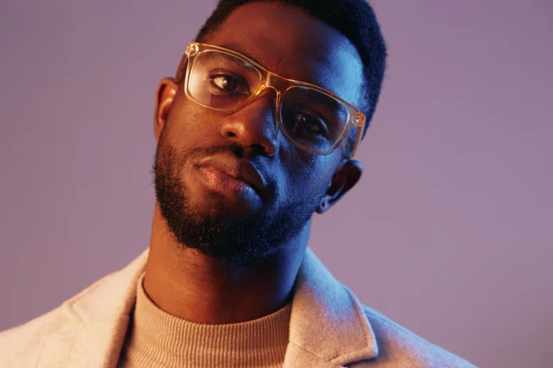 Stylish African American man with glasses showing confidence against a colorful gradient background in a fashion forward outfit